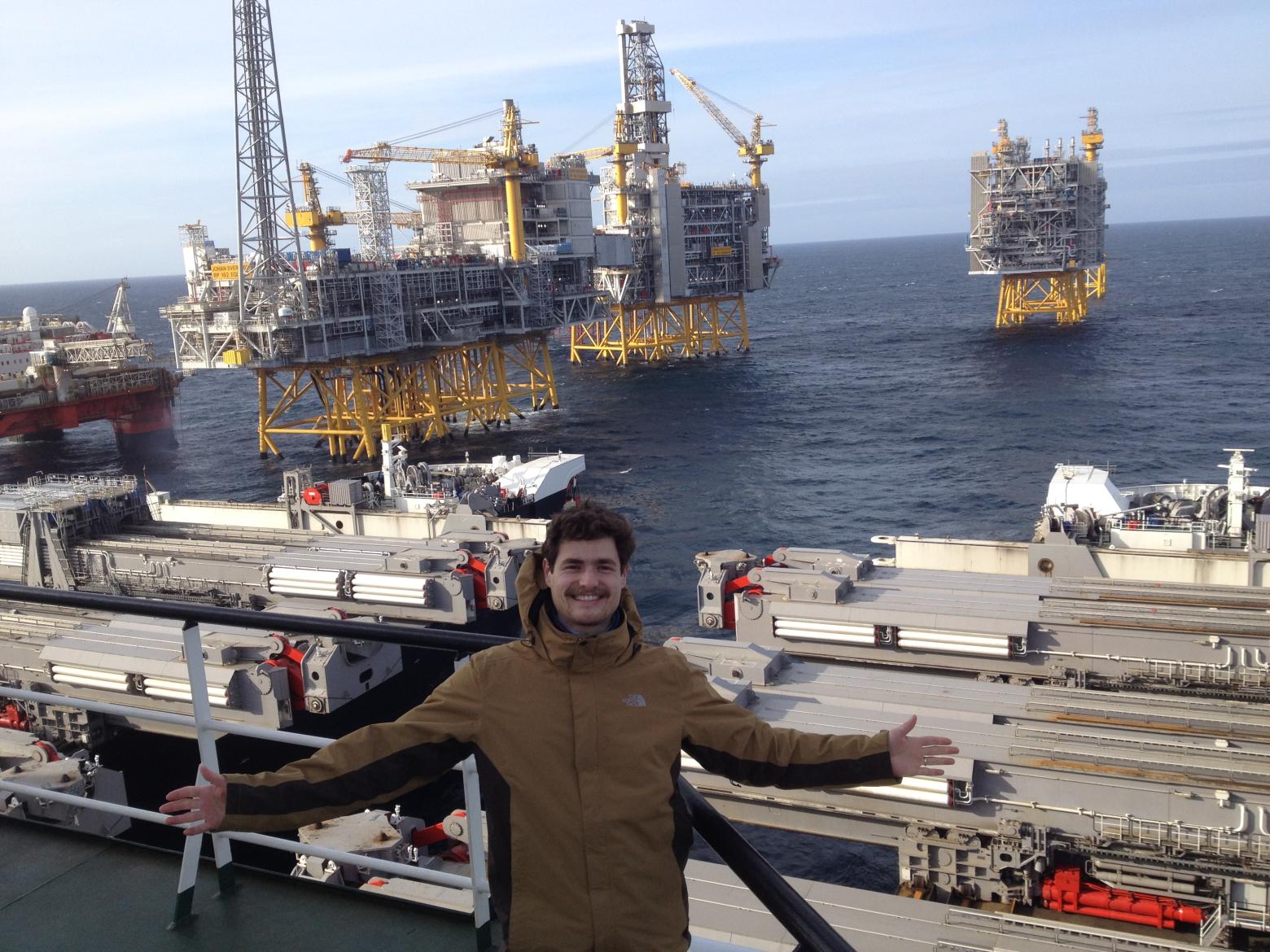Helio stands on a boat, with a large industrial structure in the background, showcasing a maritime scene.